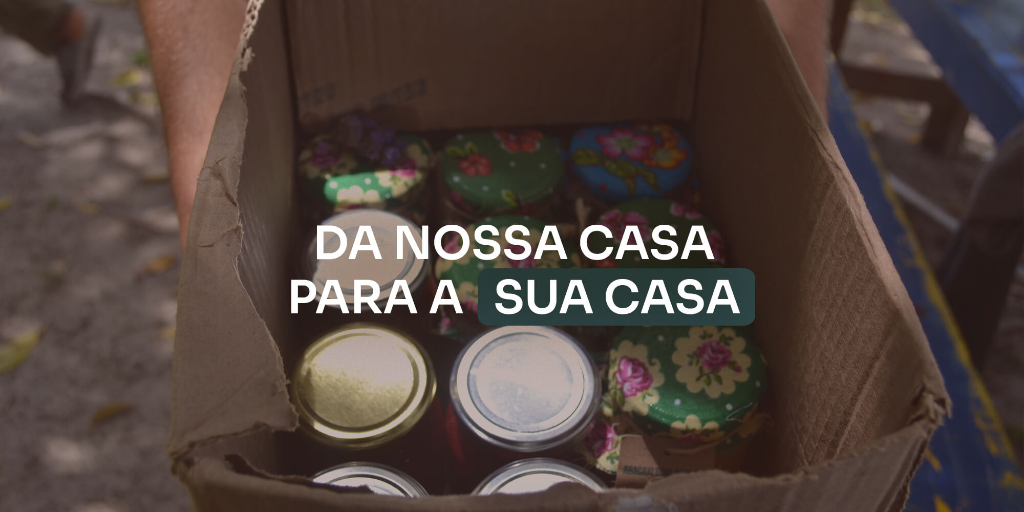 Caixa de papelão com potes de conserva artesanais dentro, com o texto "DA NOSSA CASA PARA A SUA CASA"